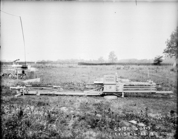 Hay presses in field. The hay press in the foreground has "I.H.C. Press" painted on the side. In the background is a fence.
