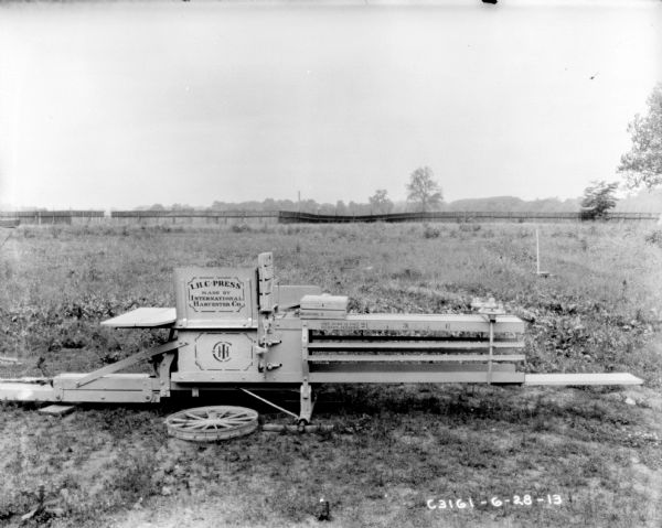 Hay press outdoors in field, with "I.H.C. Press" painted on the side. There is a wheel on the ground in front of the press, and in the background is a fence.