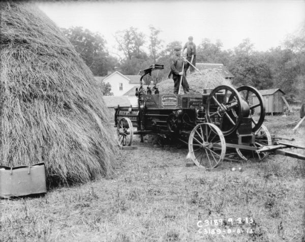 Two men using an International Harvester hay press at Springfield Works in Ohio. On the left is a large haystack. There are farm buildings in the background.