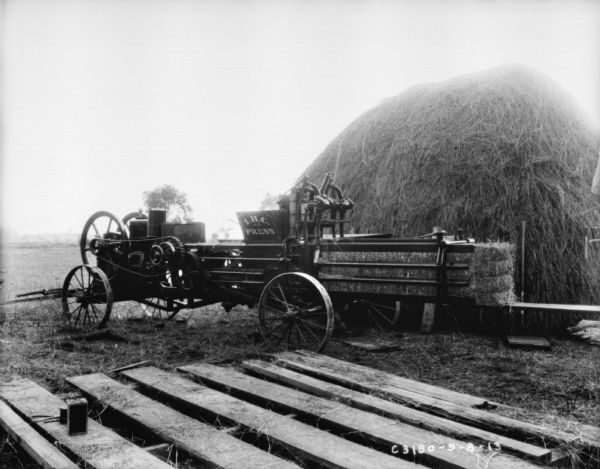 I.H.C. hay press with bales of hay in the machine. There is a pallet in the foreground. Behind the press is a large haystack.