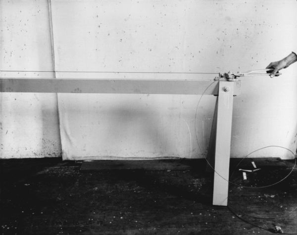 The hand of a man is holding a tool that stretches wire on a stretcher. There is a white cloth in the background as a backdrop.