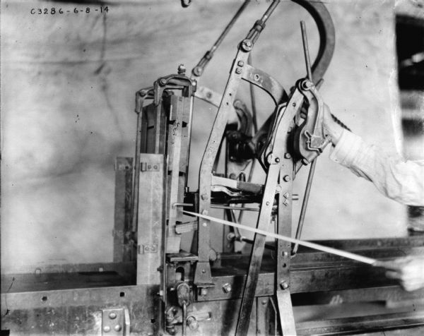 Section of hay press. From the right side can be seen a man's hands and arms, with one hand holding what may be a pointer to one section of the machine, and with the other hand holding onto a rod attached to the machine.