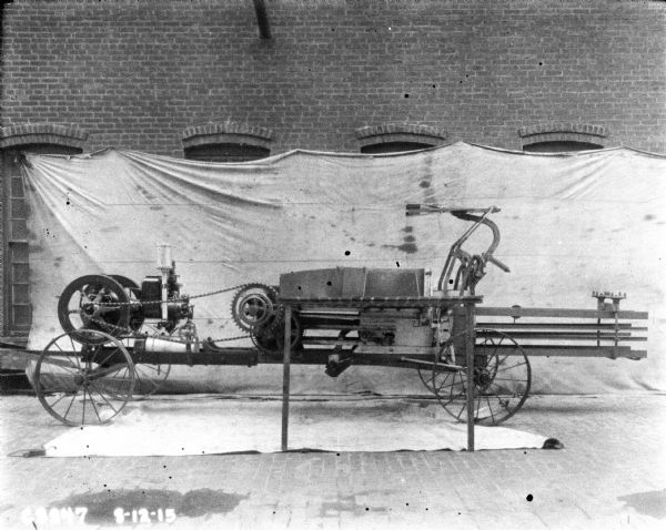 Powered hay press set up outdoors in front of a backdrop against the brick wall of a factory building.