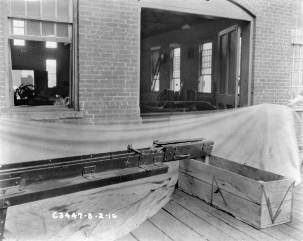 Section of hay press parts set up outdoors on a wood platform near the open door of a brick factory building. The hay press is resting on a wooden box.