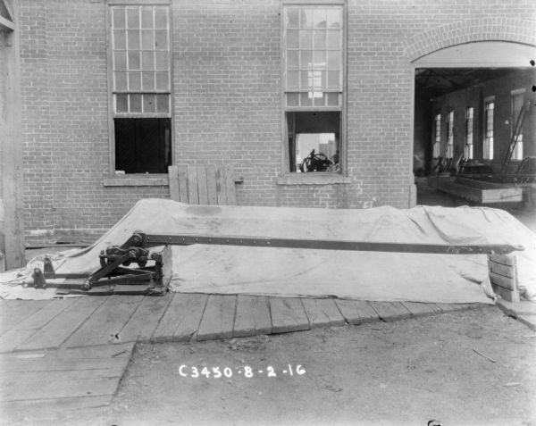 Hay press set up against a backdrop on a wood platform near the open doorway of a brick factory building. Written on the arm of the press on the right is the word: "PIN."