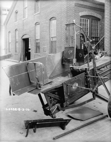 Hay press set up outdoors near a brick factory building with the number "53" between two windows. The parts on the hay press machine have been numbered, and a few parts are lying on the ground underneath.