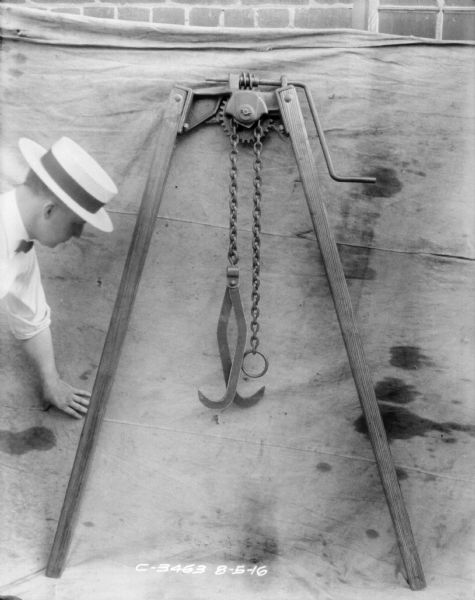 A man wearing a straw hat and a bowtie is posing with a lifting jack set up outdoors against a backdrop on a brick factory wall.