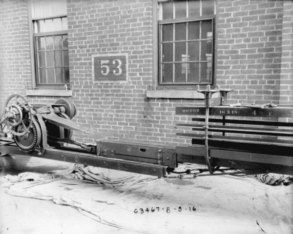 Powered hay press set up outdoors on a dropcloth near a factory building with the number "53" painted on the brick wall.