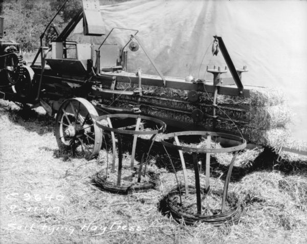 Baling operation with self tying hay press in a field. Spools of binding wire are in the foreground. There is a backdrop hanging behind the press.