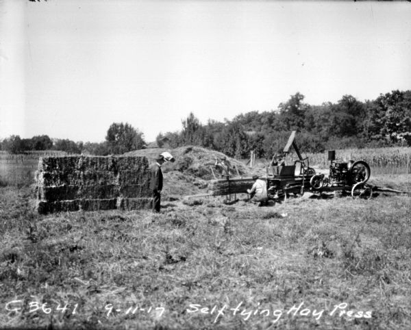 Three men are working with a self tying hay press. There is a haystack in the background. A man wearing a suit is standing and watching near a stack of hay bales on the left.