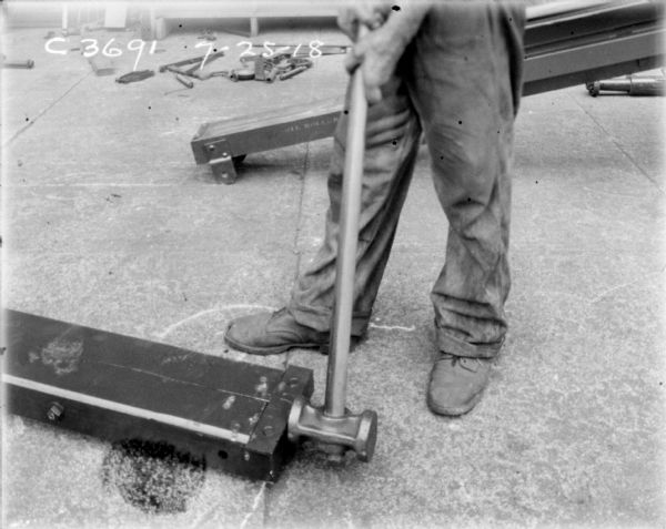 Man, (only hands and legs visible) using a hammer to make an adjustment on a piece of equipment lying on the ground.
