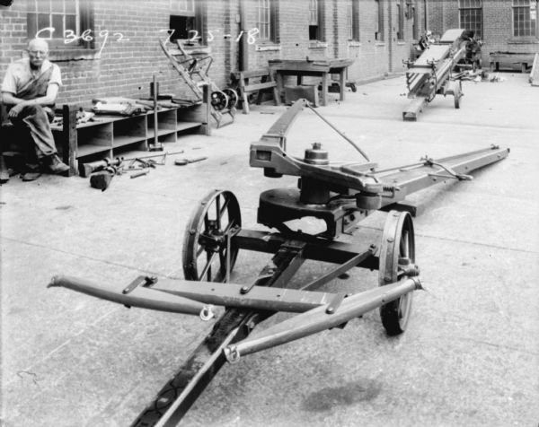 Agricultural machinery and parts set up in a yard. A male employee is sitting near the brick factory wall on the left.