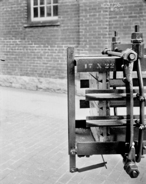 View of section of hay press outdoors near brick factory building. Numbering on press reads: "17 X 22."