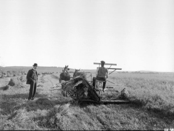 A man wearing a suit and a bowler hat is standing near a man using a horse-drawn binder in a field.