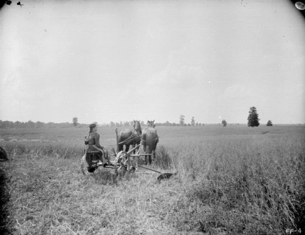 View from rear of a young girl, barefoot, sitting on a horse-drawn mower in a field holding the reins of the team of two horses.