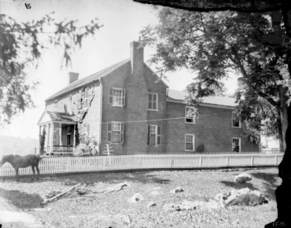 View across yard towards a large brick house surrounded by a picket fence. There is a horse in the yard on the left.
