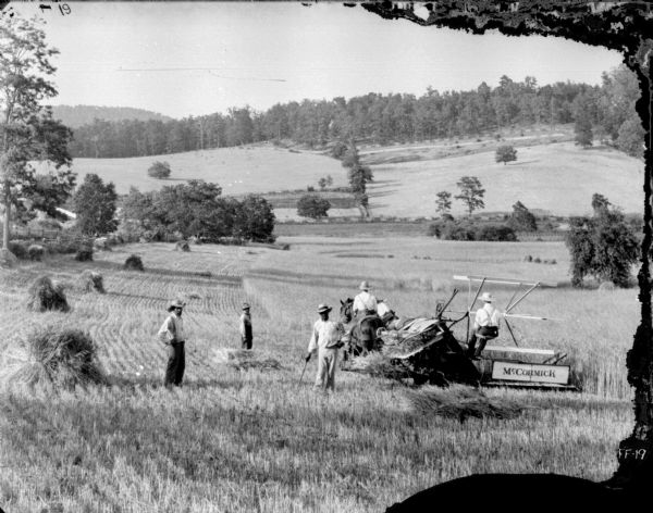 View down hill towards two men and a young boy standing in a field near two men using a horse-drawn McCormick binder. One of the men is riding one of the horses pulling the binder.