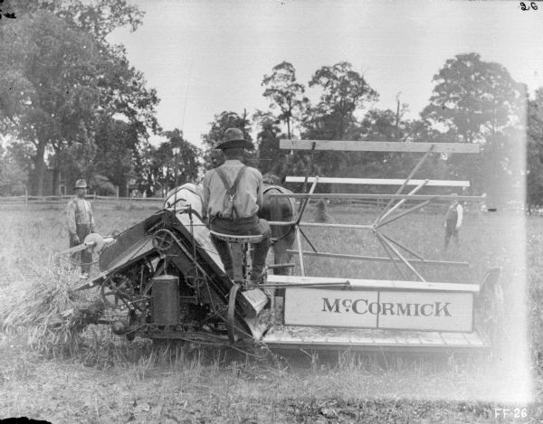 Rear view of a man using horse-drawn McCormick binder in a field. One man is standing in the background on the left, and another man is on the right. In the far background beyond a fence are trees and buildings.