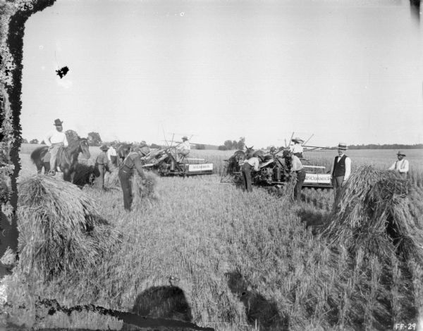 Men are working in field with shocks of grain as two men are using horse-drawn McCormick binders in a field. A man on the left is on horseback. Two other men wearing hats and bow ties are standing.