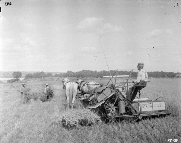 Three-quarter view from rear of a man using a horse-drawn McCormick binder in a field. In front of the horses on the left, two men are working with shocks of grain. There is a fence, trees, and what may be a river in the far background.