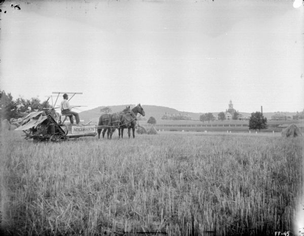 On the left a man is posing with a horse-drawn McCormick binder in a field. In the distance across fields and fences is a large estate.