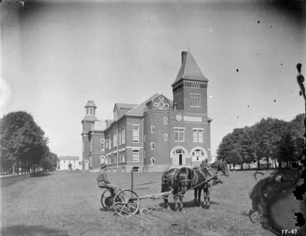 Right side view of a man on horse-drawn binder on the lawn of an estate. A large brick building is in the background. On the left is a fence-lined road with trees.