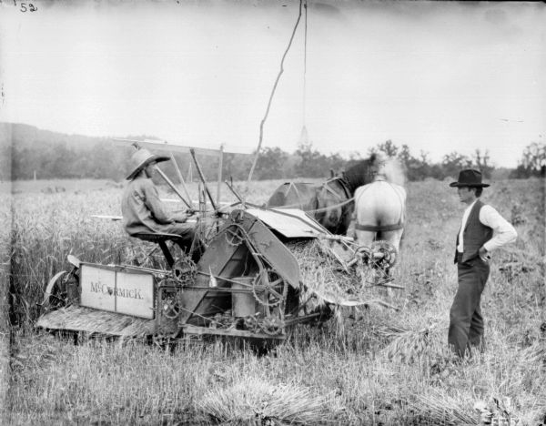 View towards a man on the left sitting on a horse-drawn McCormick binder. A man wearing a hat and vest is standing on the right.