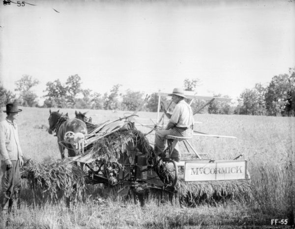 Rear view of a man using a horse-drawn McCormick binder in a field. A man is standing on the left near the binder.