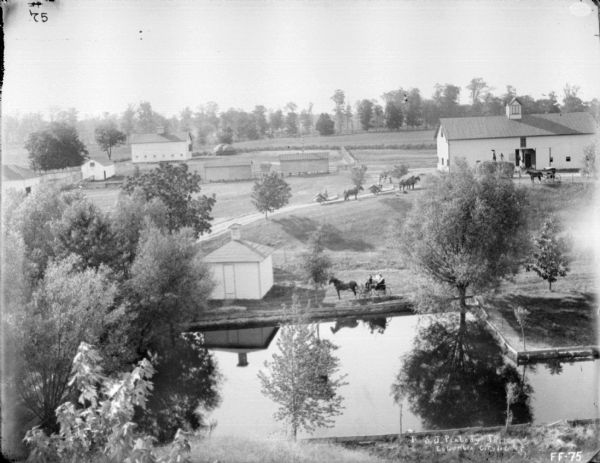 Elevated view from hill looking down at a lagoon and farm buildings. People are posing in a horse-drawn carriage on the far shoreline. More people are on horse-drawn binders near farm buildings, roads, and a large barn.