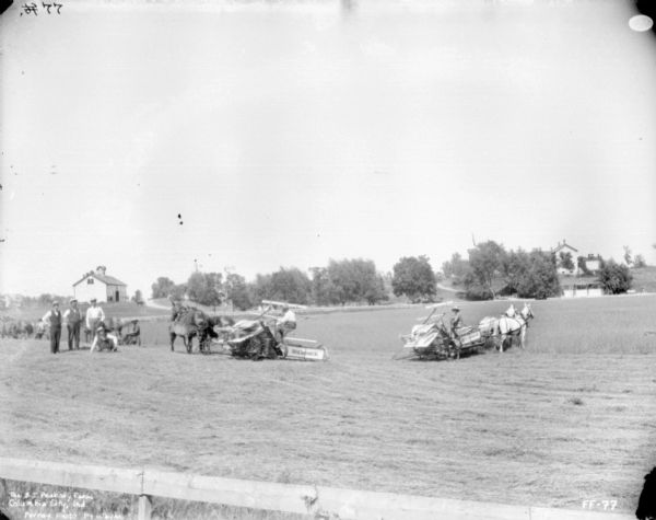 Four men wearing vests are posing in a field along with four men on horse-drawn mowers and McCormick binders. In the background are farm buildings.