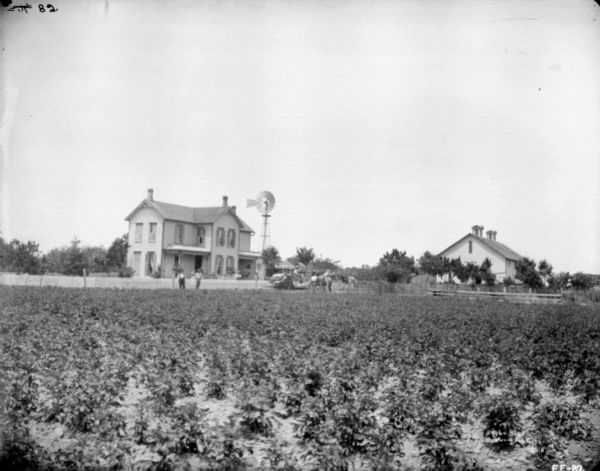 View across field towards a group of people posing on a farm. Men are standing near a fence in front of a farmhouse and a windmill. In the center is a man on a horse-drawn binder. On the right is a barn.