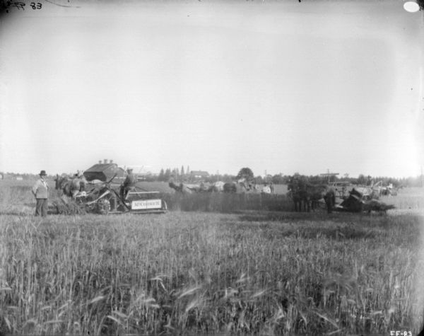 View across field towards a group of men standing near other men using horse-drawn McCormick binders. In the background are farm buildings.