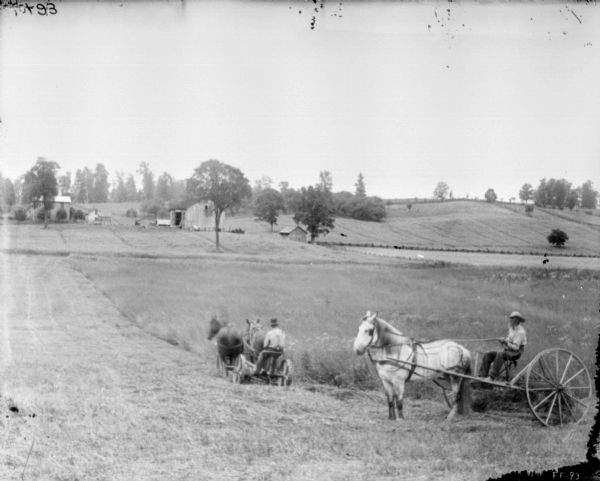 View down slope towards a man using a horse-drawn mower in a field, and another man, on the right, with a horse-drawn dump rake. The horse pulling the rake is wearing a fly-net. Farmhouse and farm buildings are in the background.