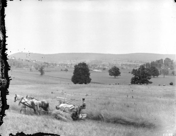 View down hill towards a man using a horse-drawn binder in a field. In the valley in the distance is a town.
