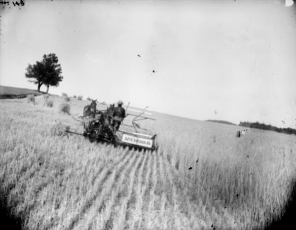 View across harvested field towards a man using a horse-drawn McCormick mower on the slope of a hill.