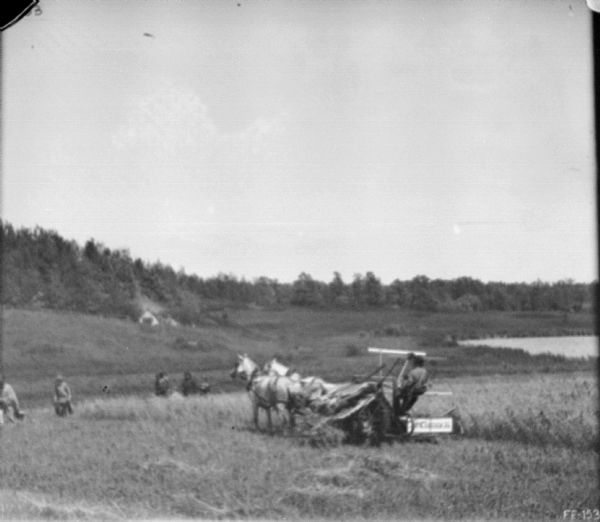 Men are working in a field near a man using a horse-drawn McCormick binder. People are watching at the edge of the field in the background. On a hill above them near trees is a tipi. There is a body of water on the far right.