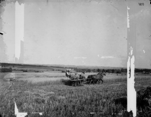 View across harvested field towards three men working with a horse-drawn binder in a field. In the distance are more fields and low hills.