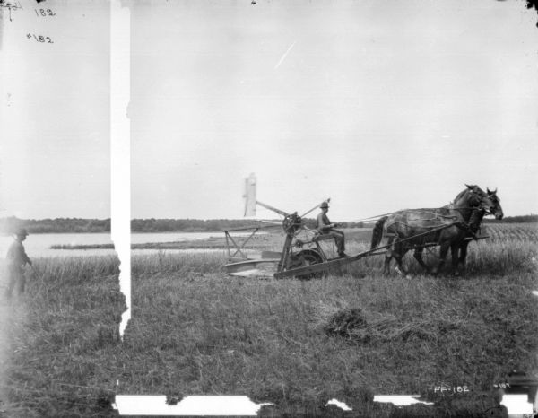 Right side view of a man using a horse-drawn reaper in a field. Behind him on the left is a man. In the background is a lake.
