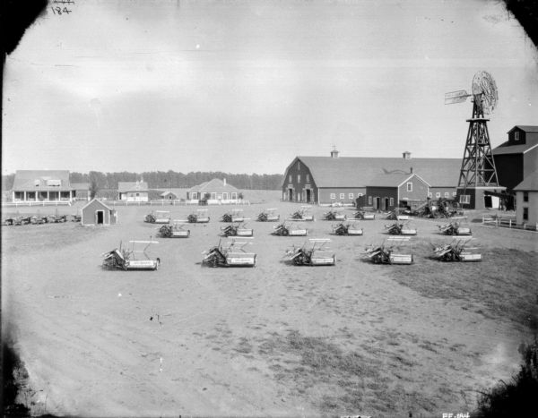 Elevated view of approximately 19 McCormick horse-drawn binders in a barnyard of a very large farm, or of a dealership. There are buildings behind a fence in the background. Large barns, and a windmill with the name Halladay on it, are on the right.