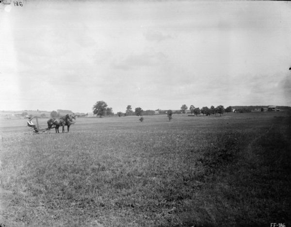View across field towards a man on a horse-drawn mower with the rake in the raised position. In the far background are trees and farm buildings.