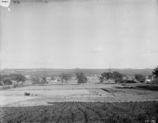 View down hill towards a man using a horse-drawn binder in a field. In the distance is a valley and hills with farm buildings.