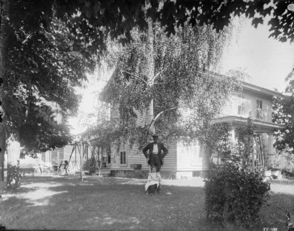 View across shady lawn towards a man standing behind a girl sitting in a chair. Behind them is a house. On the right is a lawn swing, and beyond is a woman sitting in a horse-drawn buggy.