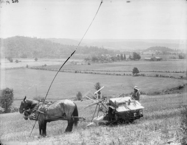 Left side view of a man on a horse-drawn binder on the side of a hill in a field. The field slopes down to a valley with more fields, and farm buildings among trees. In the distance are tree-covered hills.