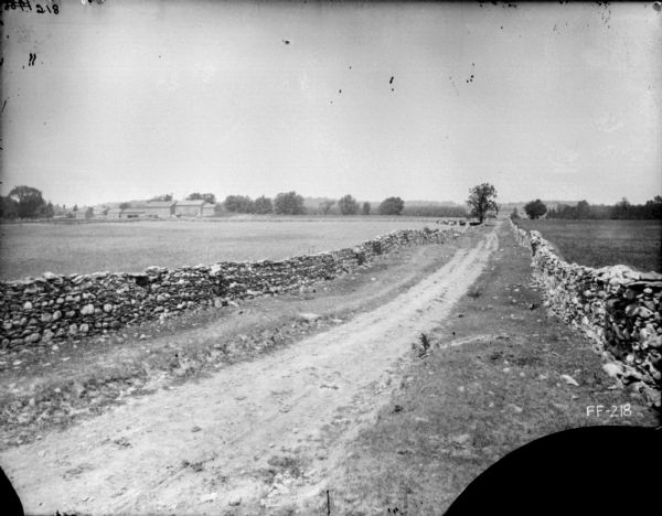 View down unpaved road, with stone walls along the sides of the fields on the left and right. There are farm buildings in the background on the left.