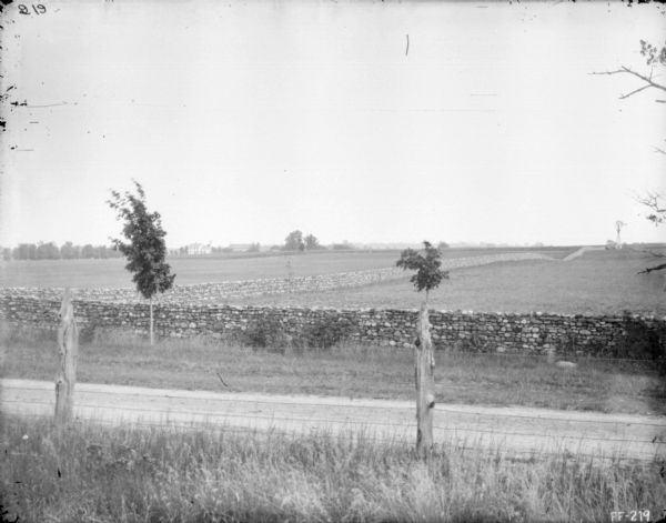 View across road towards stone walls in a field. Farm buildings are in the far background on the left. There is a windmill in the field on the right.