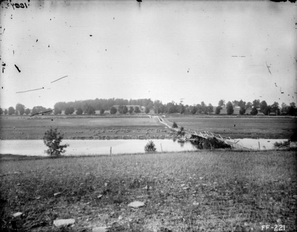 View across field towards a river with a bridge on the right. On the far side of the river is a flock of animals. Farther in the distance are farm buildings and trees.