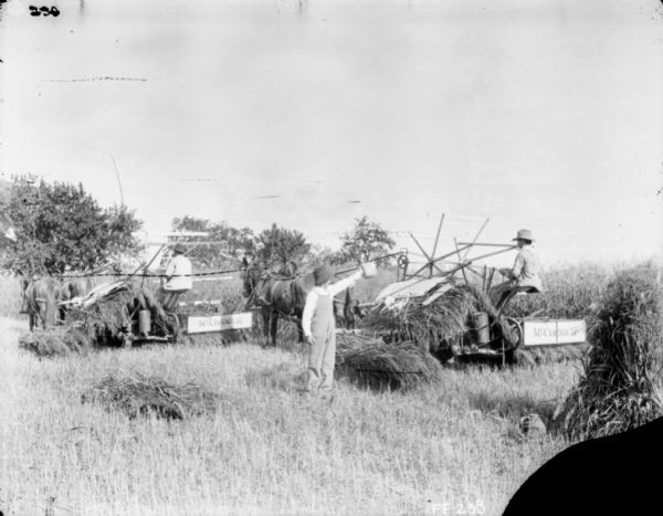 Two men are using horse-drawn McCormick mowers in a field. A young boy is standing in the center foreground holding a roll of twine.