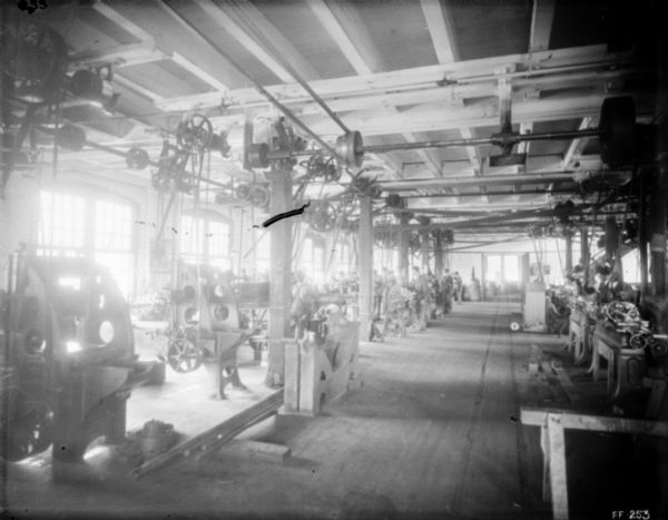 Interior view of the metal finishing room. Machines are driven by belts on the ceiling. Men are working at machines in the background.