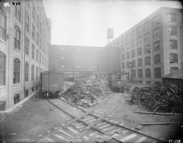 View of factory yard, with two railroad tracks crossing each other in the center, and piled between the tracks are scrap metal and lumber. Railroad cars sit on both sets of tracks. There is a brick factory building on the left and right, connected by another building in the background.