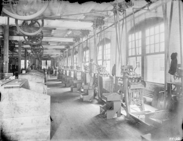 Interior view of assembly line in a factory. One man is working on a machine near the row of windows on the right.